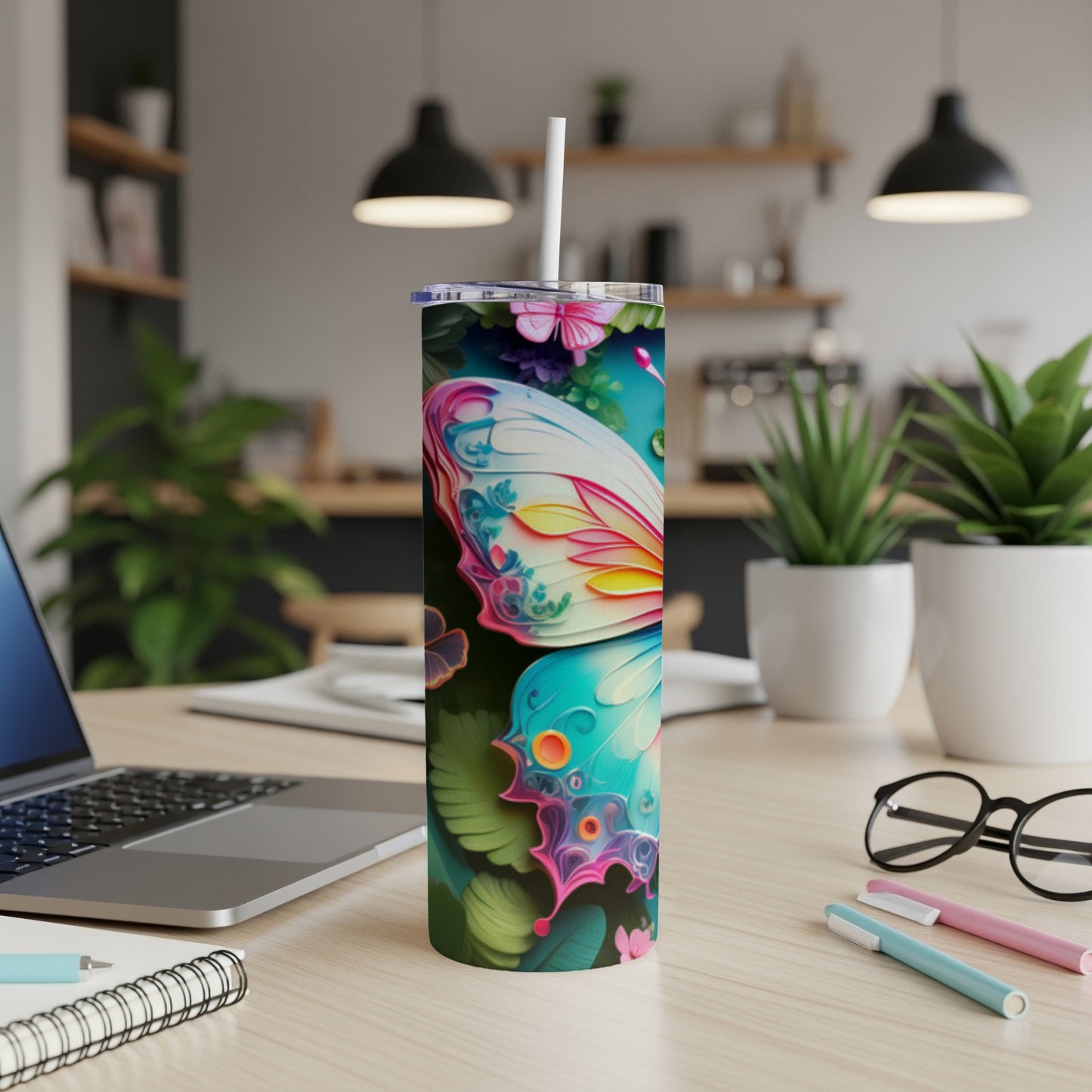 Butterfly tumbler with straw on desk near laptop and pens.