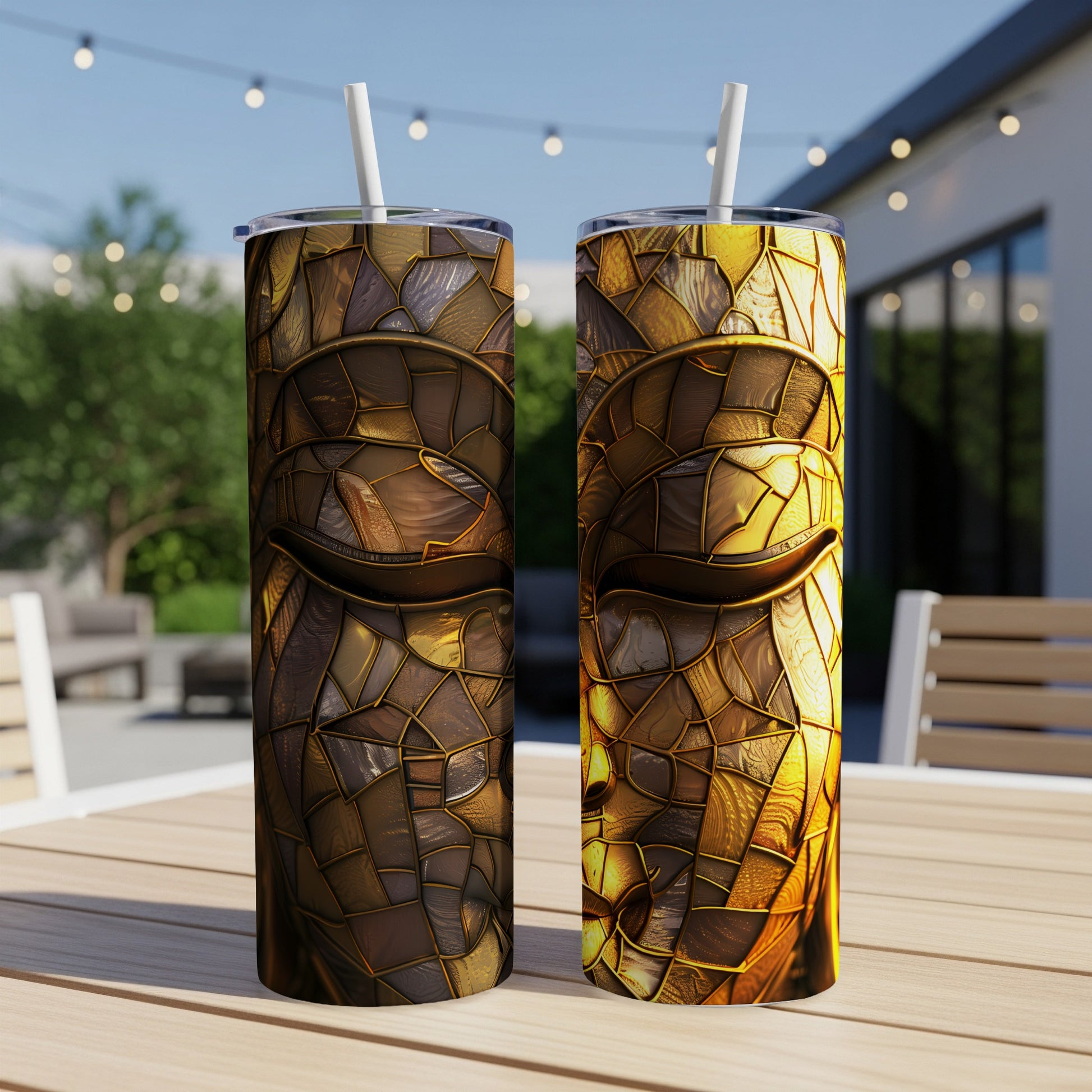 Two textured gold Buddha face tumblers with straws on an outdoor table.