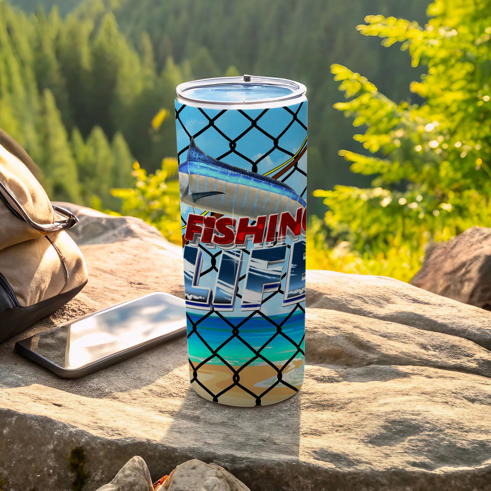 Fishing Life tumbler with a marlin graphic on a rock by a backpack and phone.