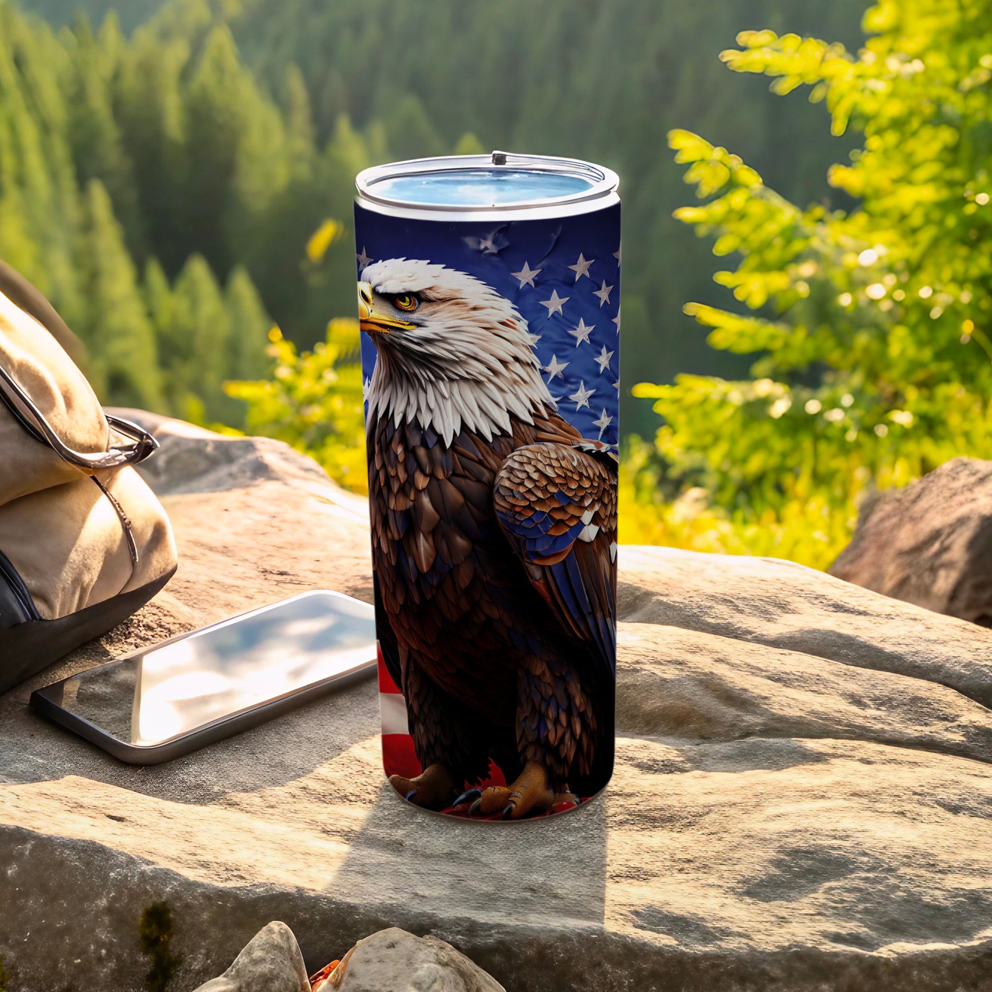 American flag tumbler with eagle design sits on rocks near a backpack and phone.