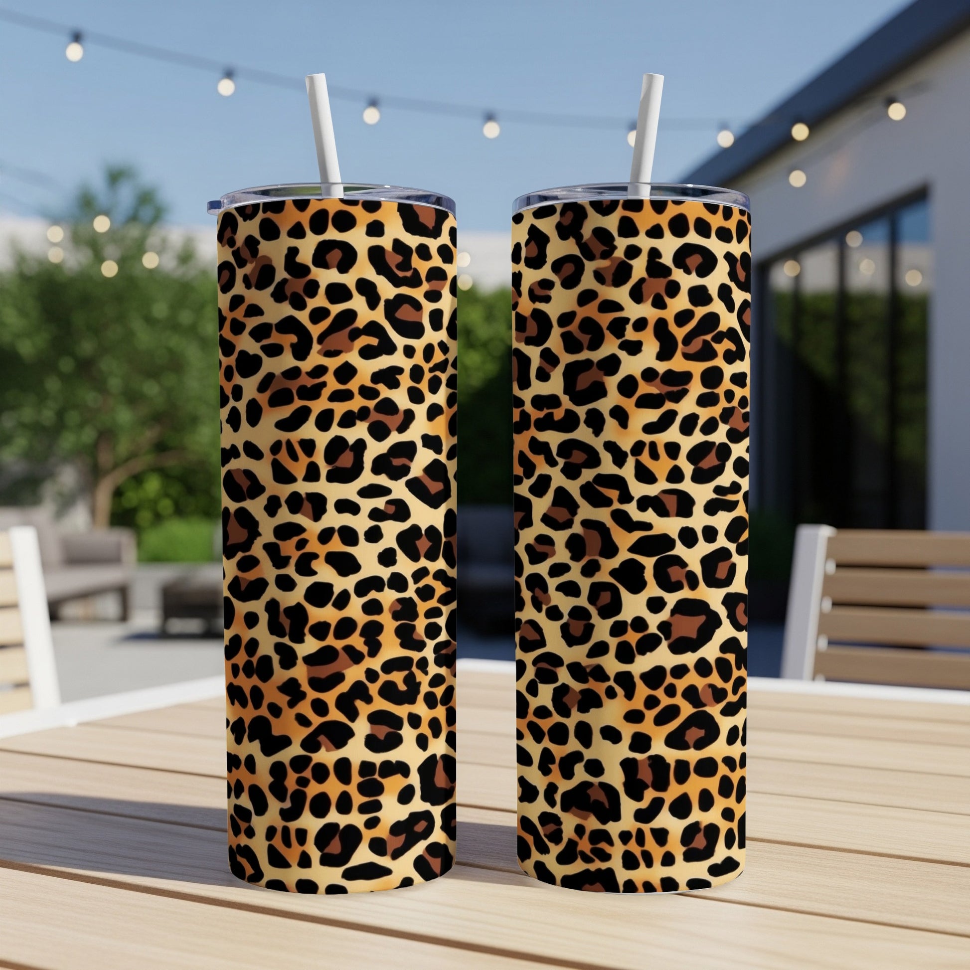 Two tall leopard print tumblers with straws on a wooden table outdoors.