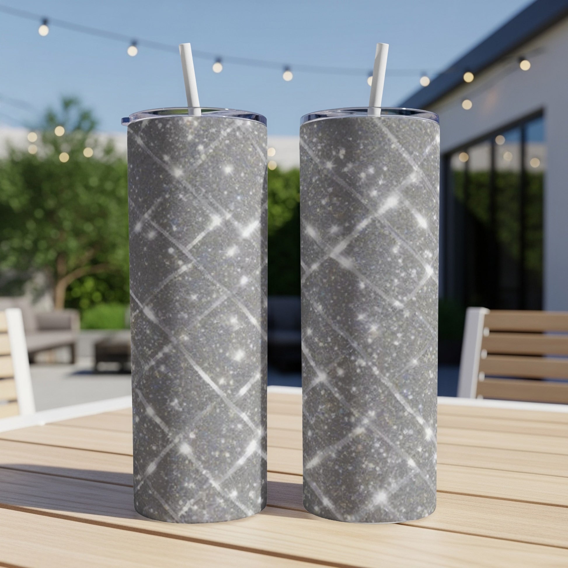 Two tall glitter tumblers with straws sit on a wooden table outdoors.
