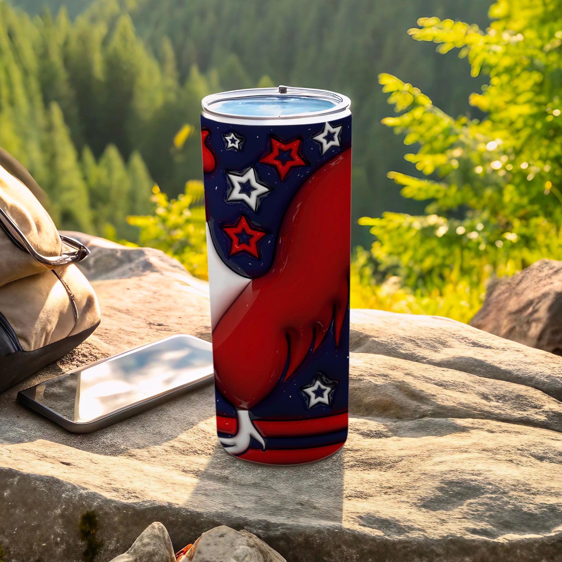 Red, white, and blue patriotic tumbler with stars on rocks with backpack and phone.