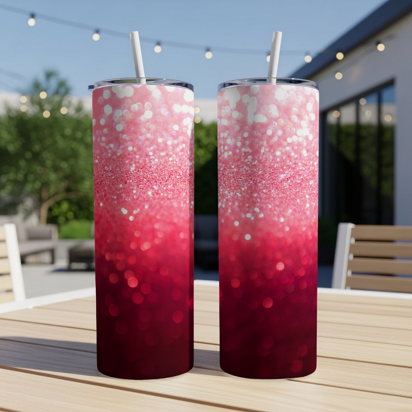 Two tall, pink to red glitter tumblers with straws on a wooden table.
