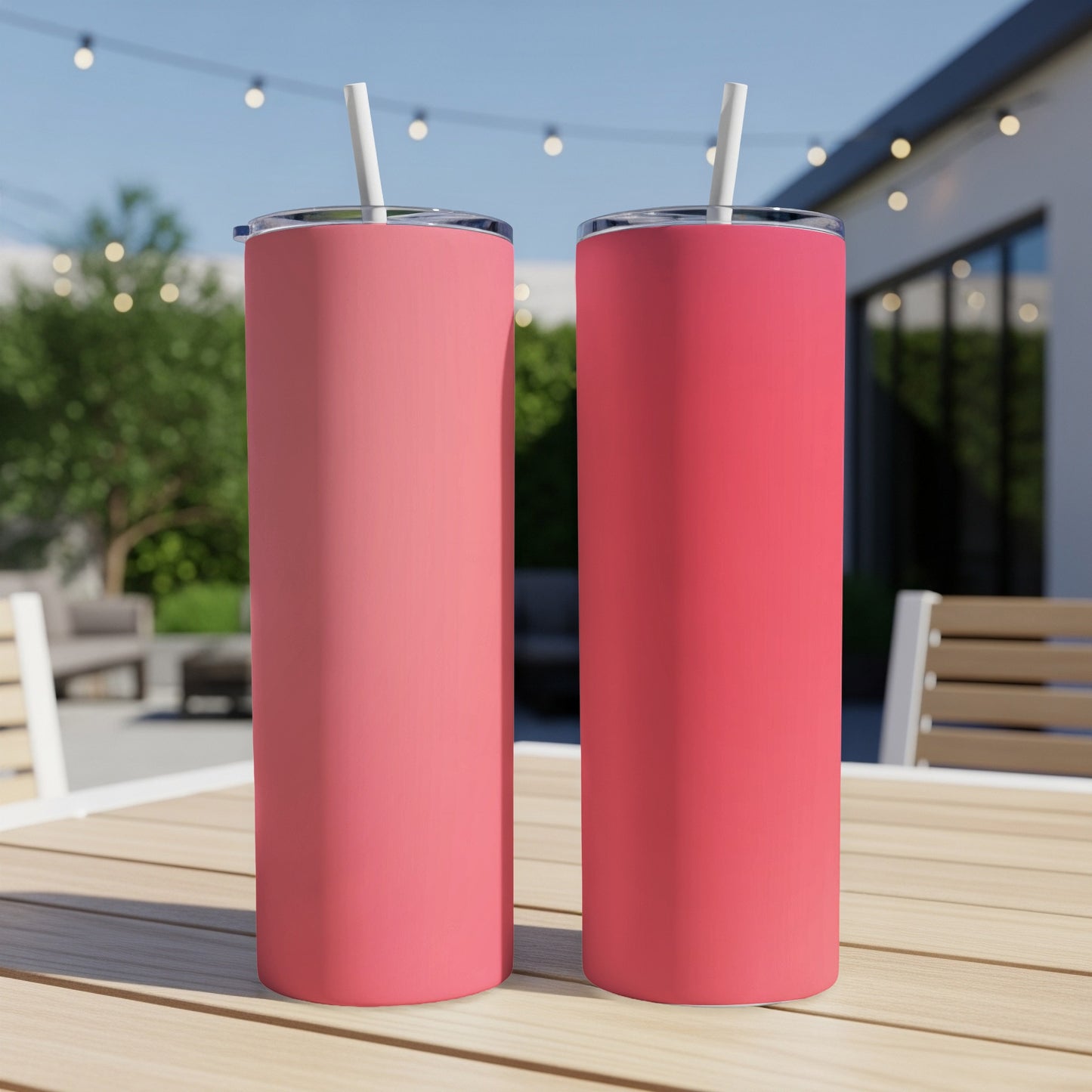 Two pink stainless steel tumblers with straws on a wooden table outdoors.