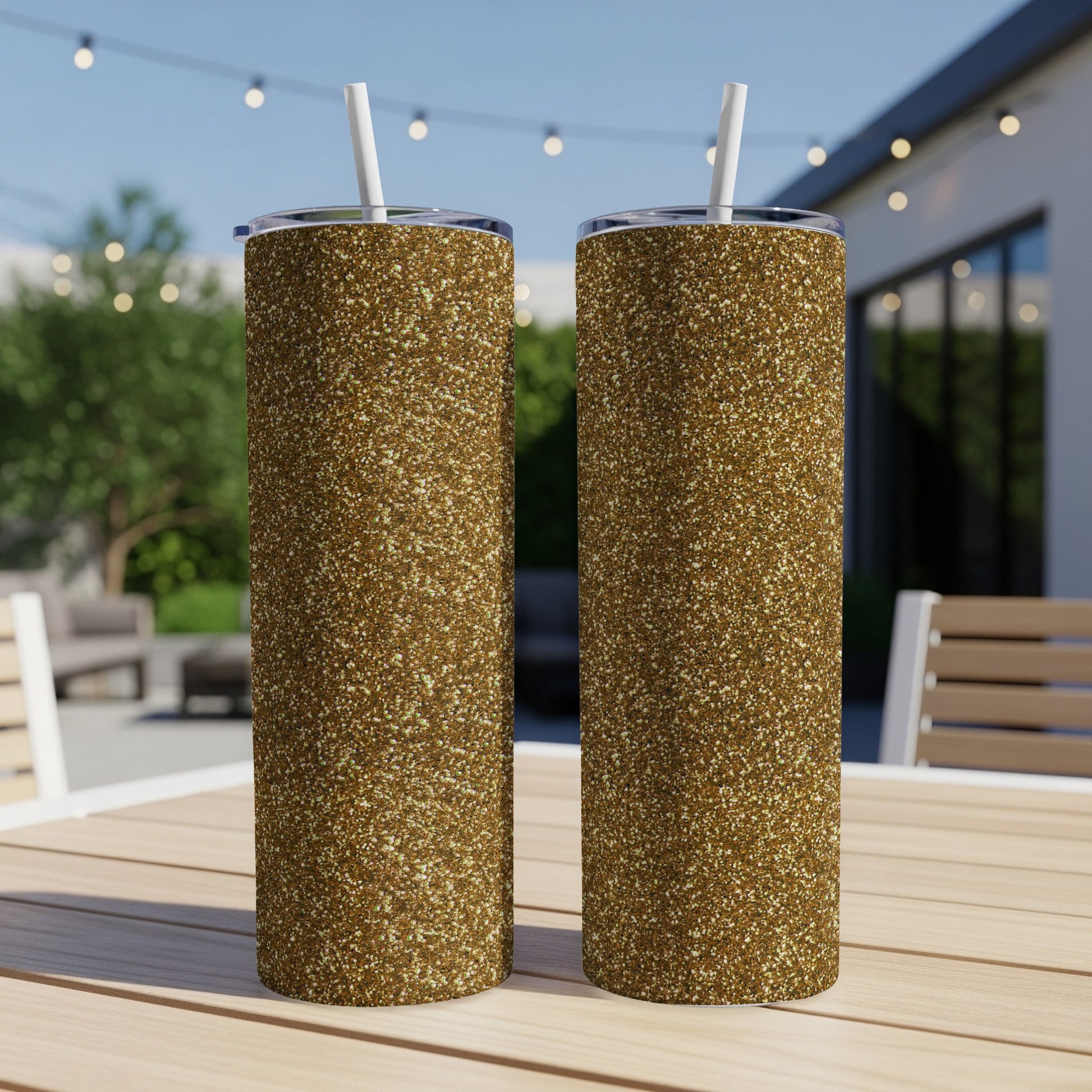 Two gold glitter tumblers with white straws sit on a wooden table outdoors.