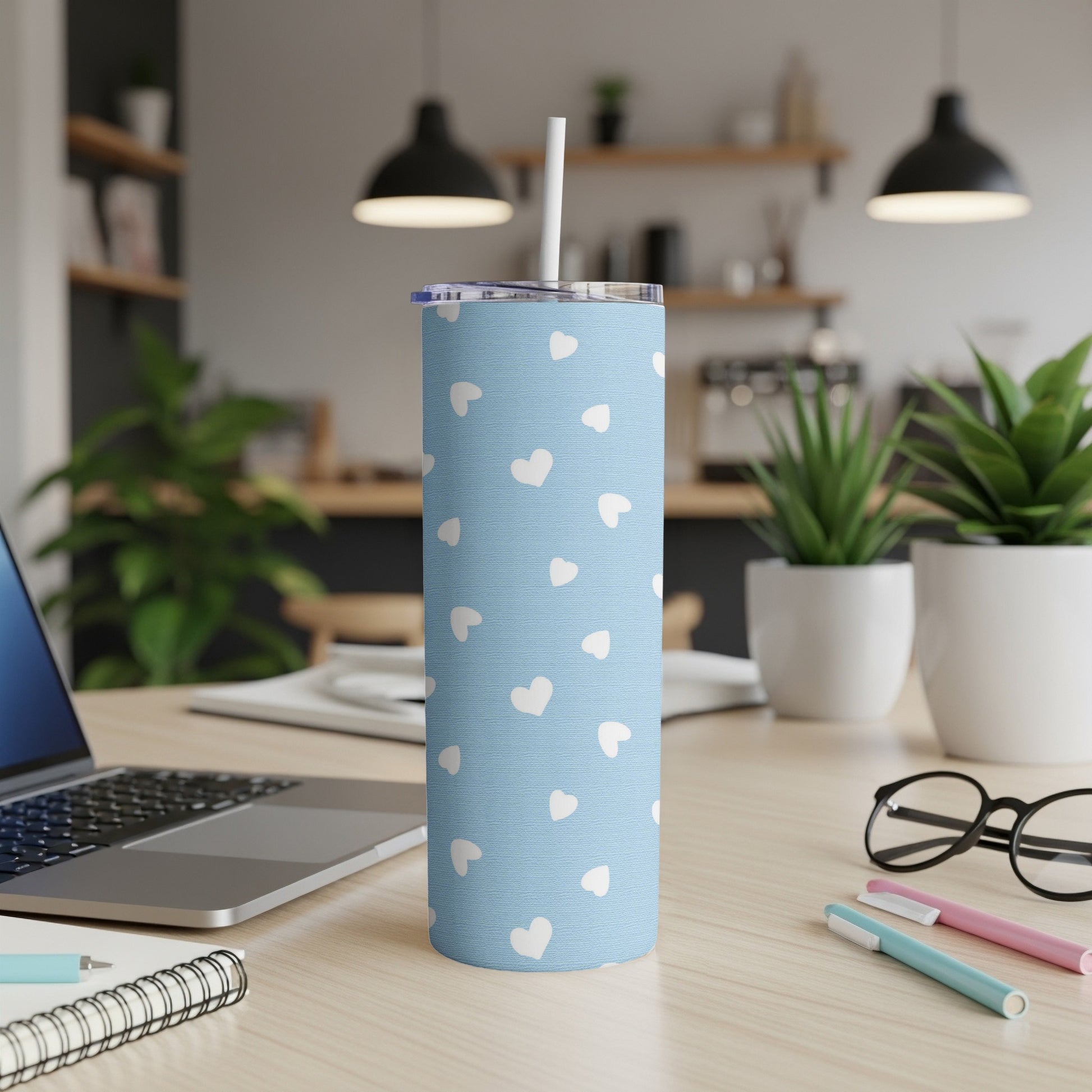 Blue tumbler with white hearts pattern, straw, and lid on a desk.