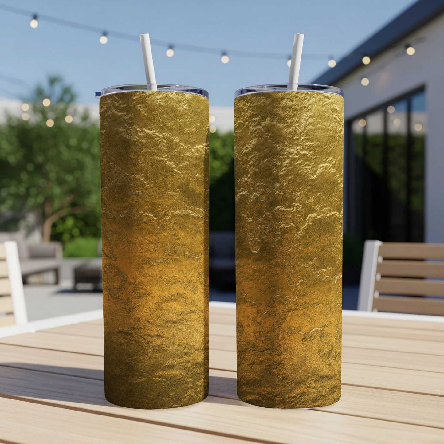 Two golden textured tumblers with straws sit on a wooden table outdoors.