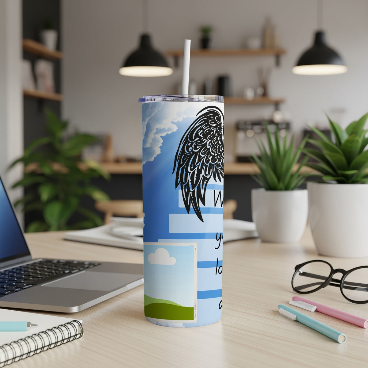 Tumbler with black wings, clouds, and text reading "When you're lost" on a desk.