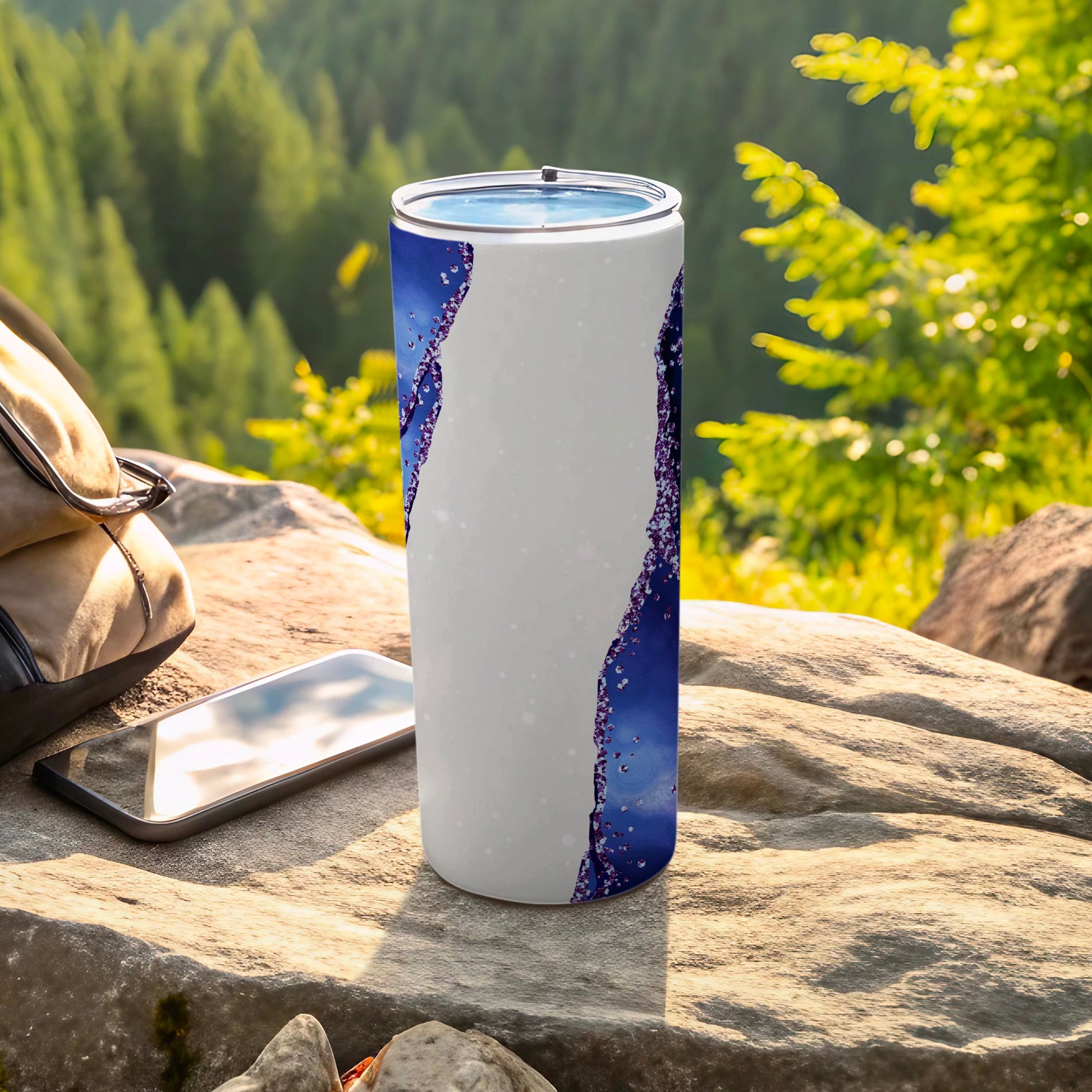 Tall white tumbler with blue sparkling water pattern, next to phone and backpack.
