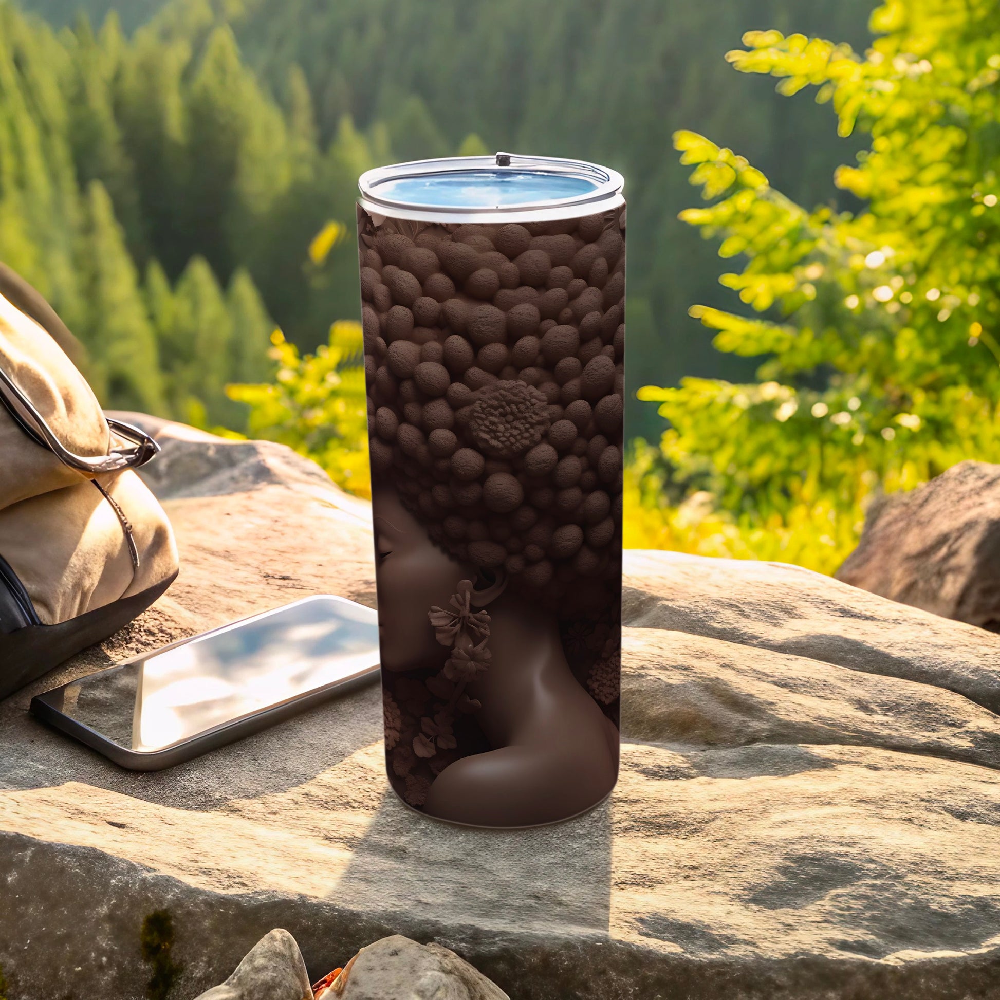 Brown tumbler with floral, profile design, on rocks with phone and bag.