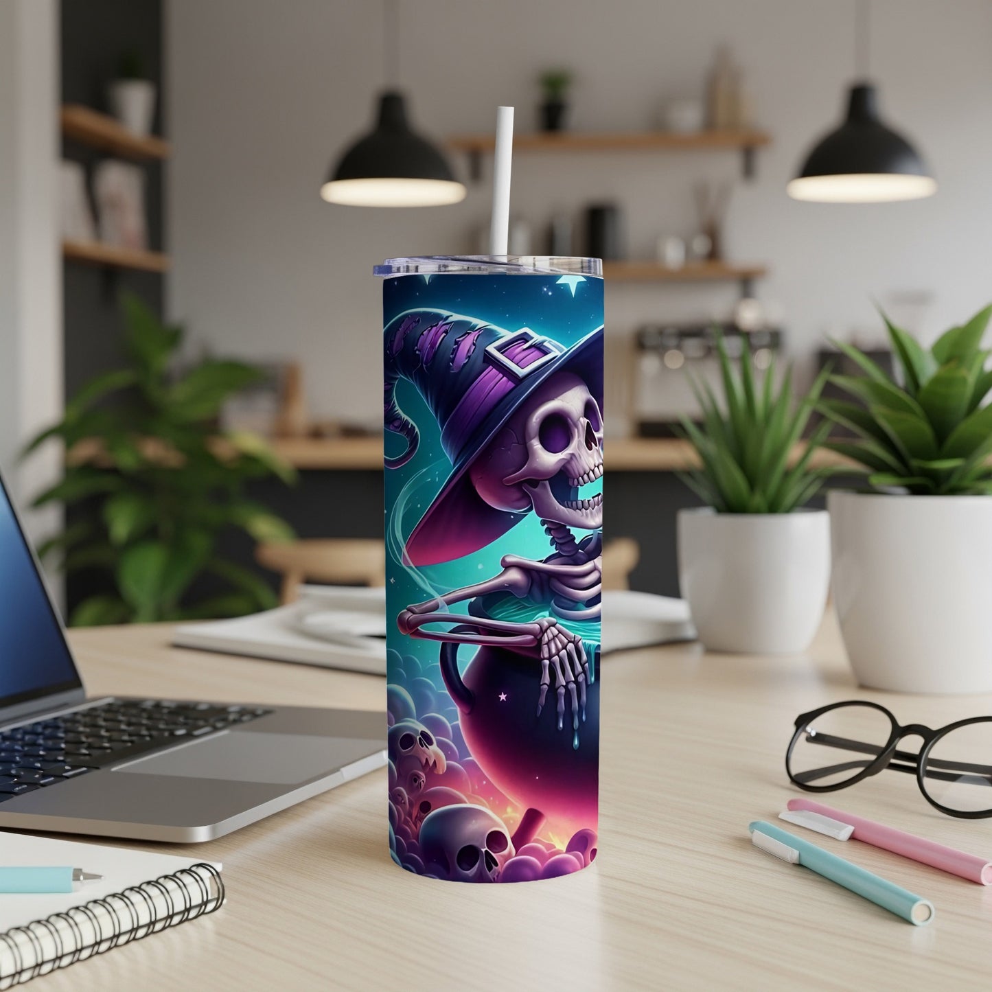 Tumbler with skeleton witch, cauldron, and skulls graphic on a desk.