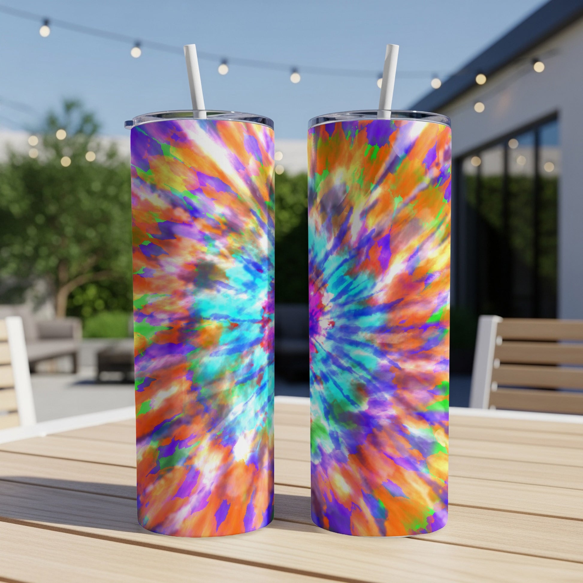 Two tie-dye tumblers with white straws on a wooden table outdoors.