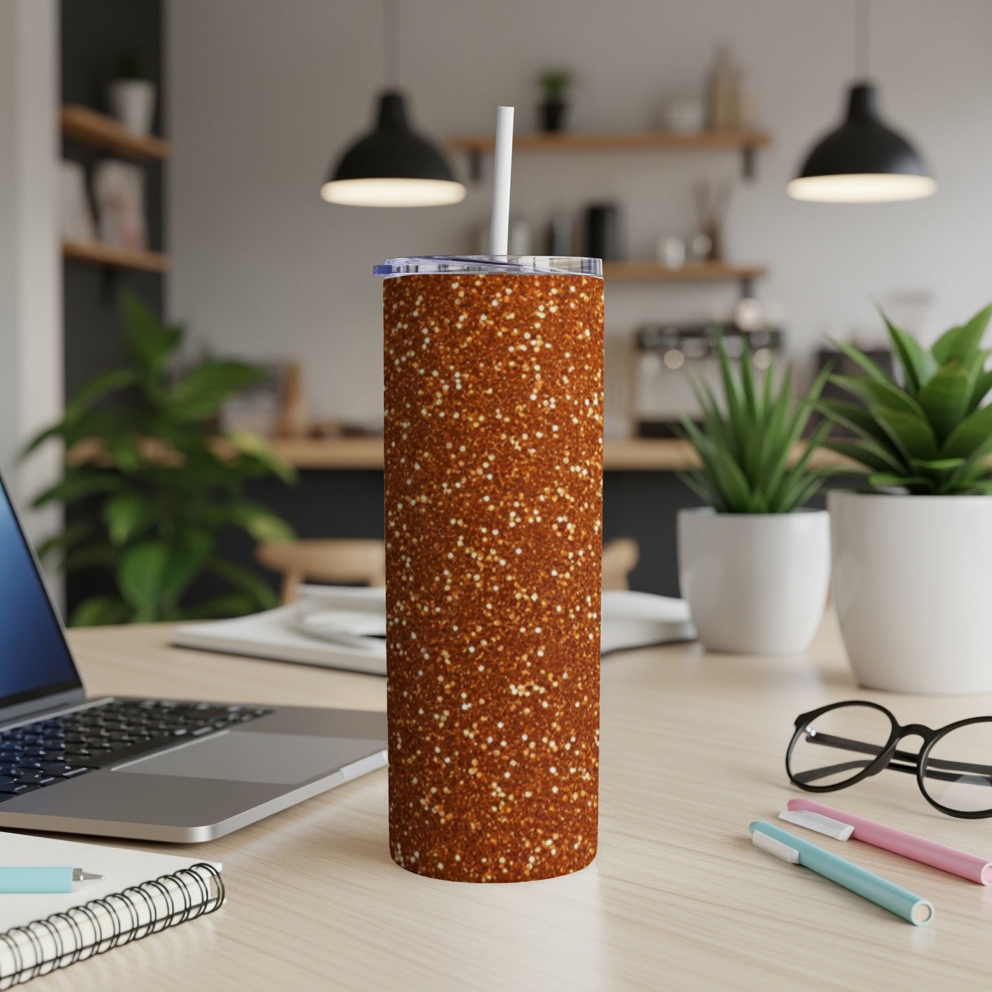 Glittery copper tumbler with a white straw on a desk with a laptop and plants.