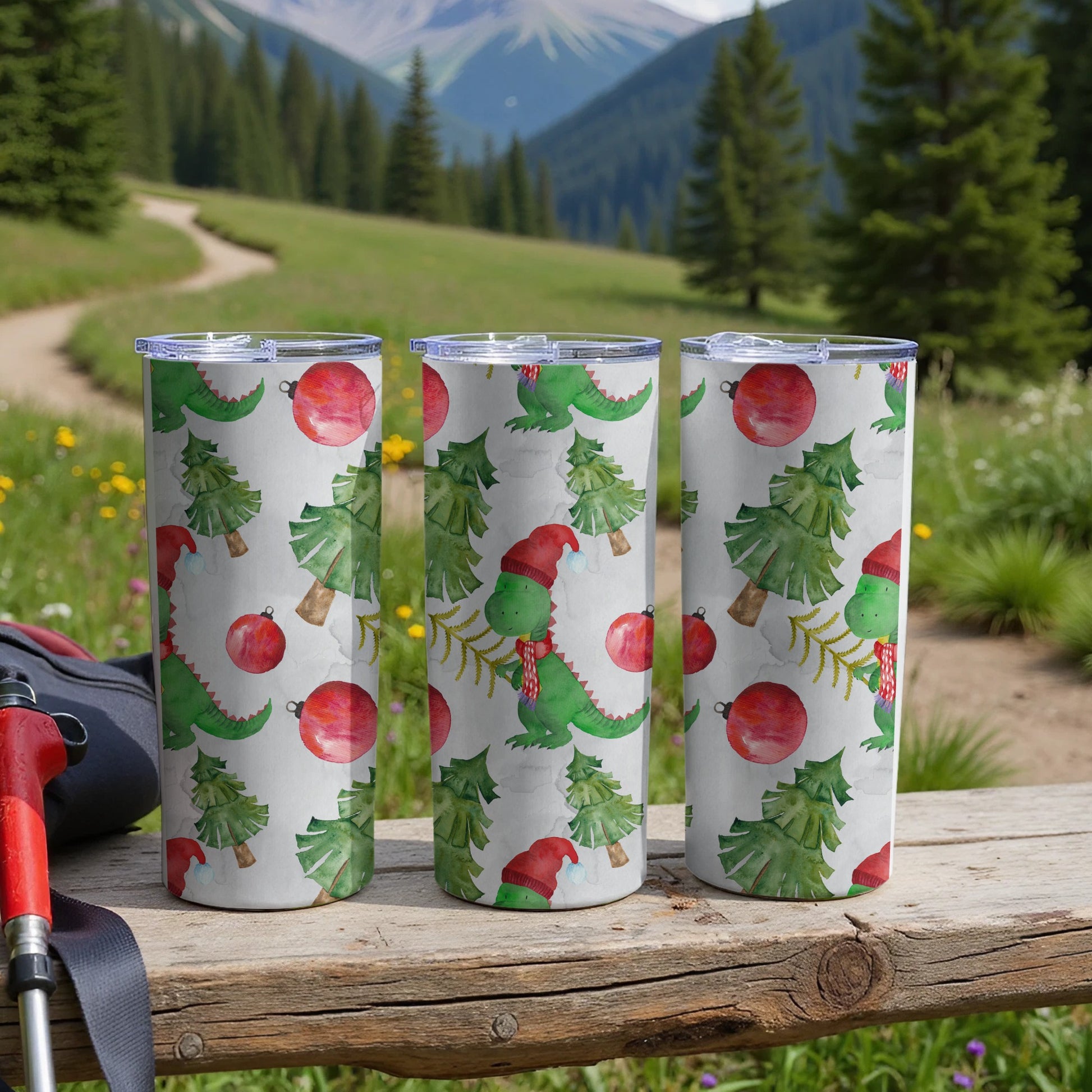 Three tumblers with a Christmas dinosaur and tree pattern sit on a wooden surface.