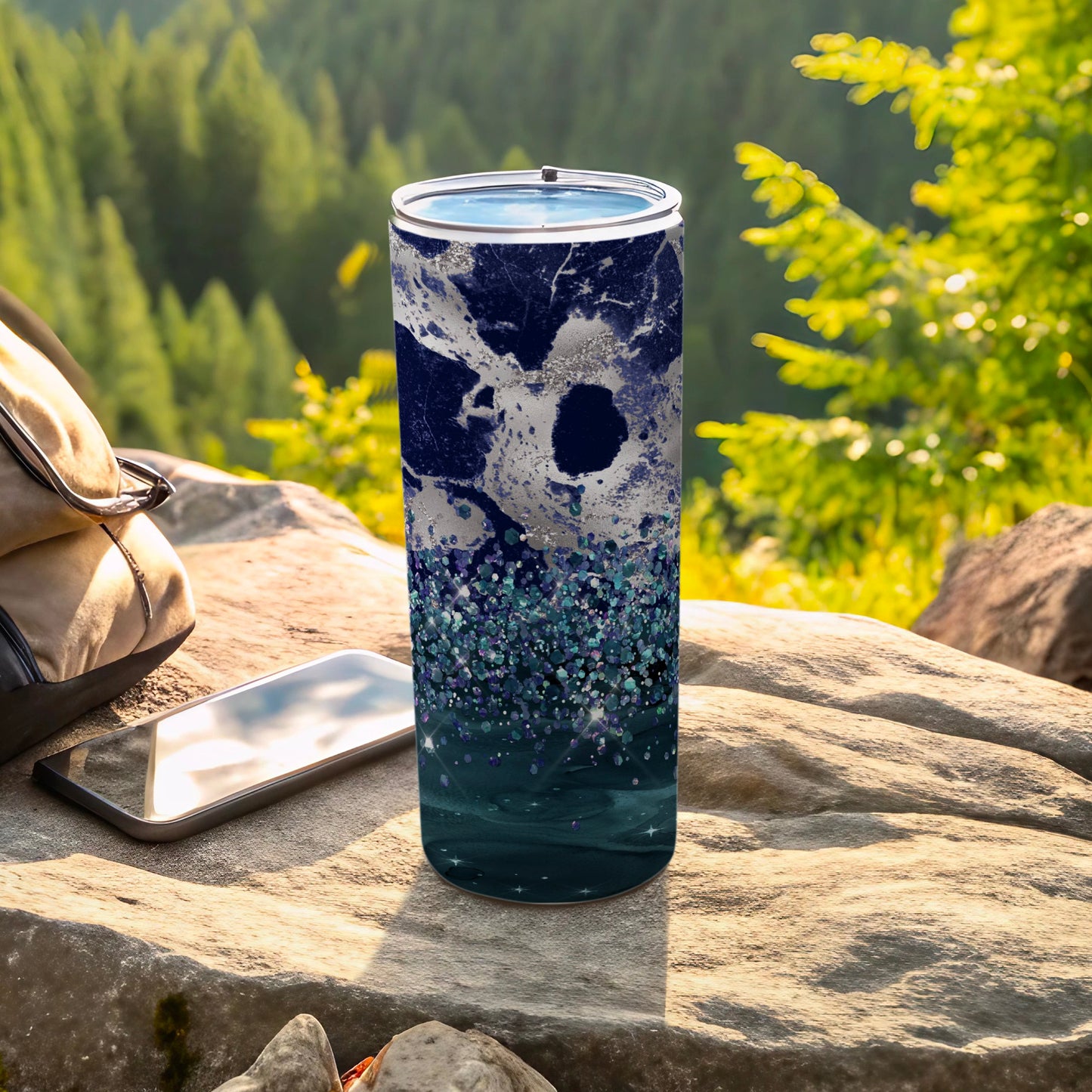 Blue marble and glitter tumbler with lid on rock near phone and backpack.