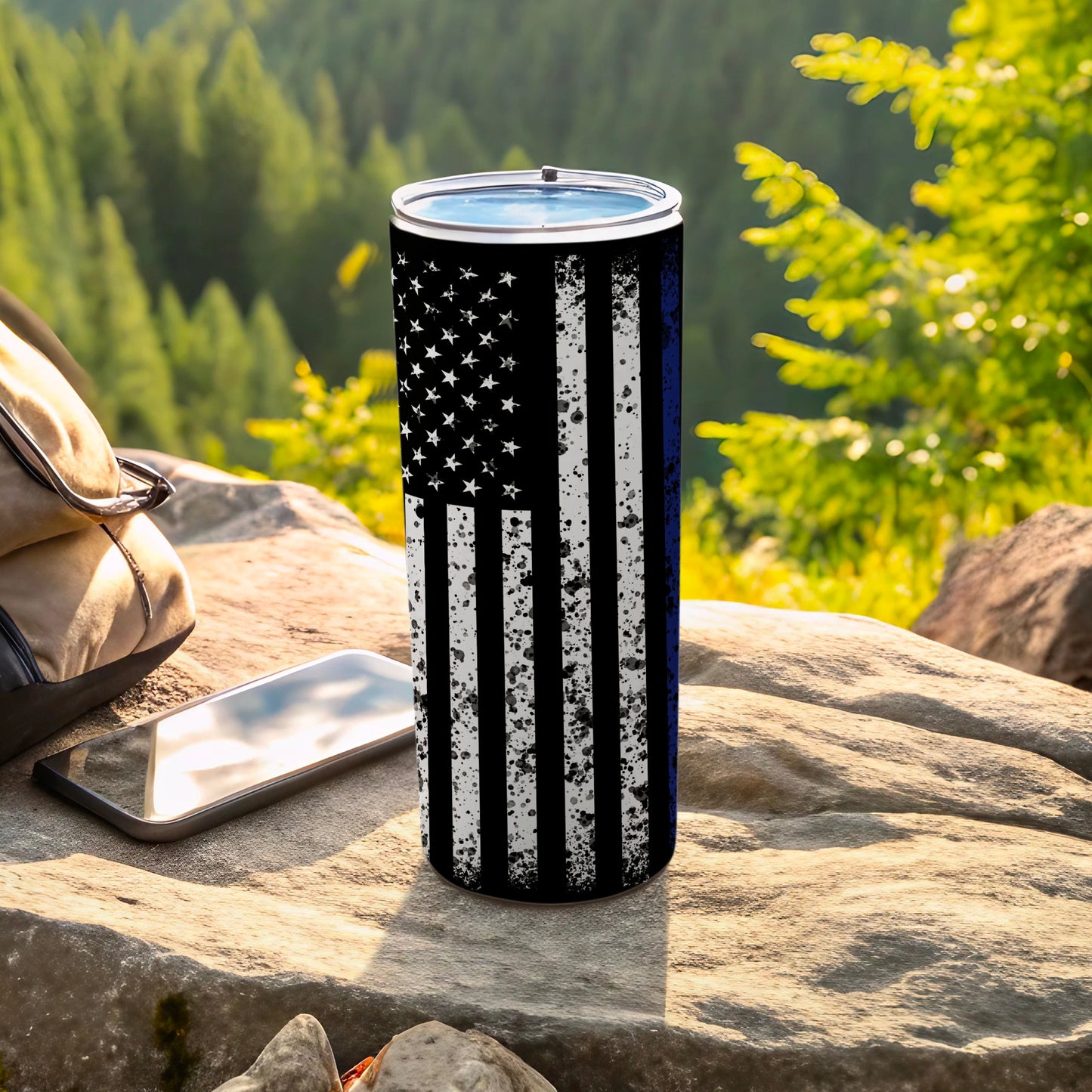 Thin blue line American flag tumbler on rock with backpack and phone.