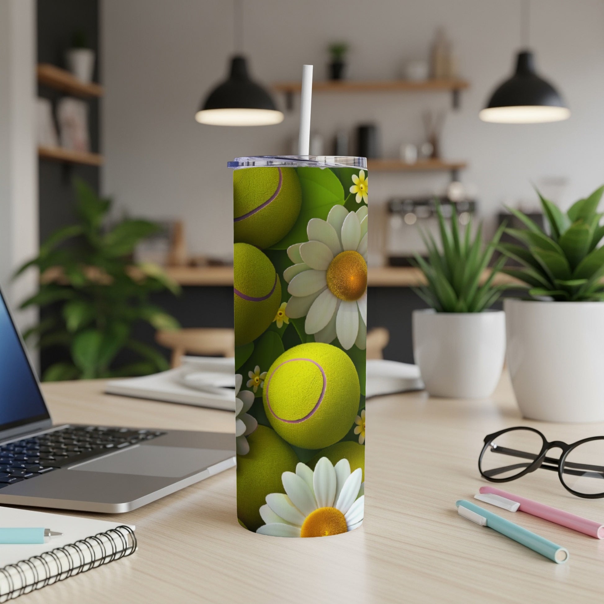 Tennis balls and daisies wrap around a white skinny tumbler on a desk.