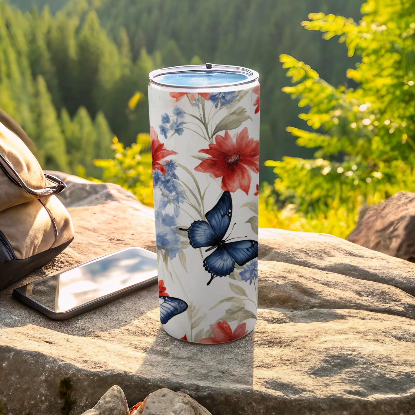 Floral and butterfly pattern tumbler on rock with phone and backpack.