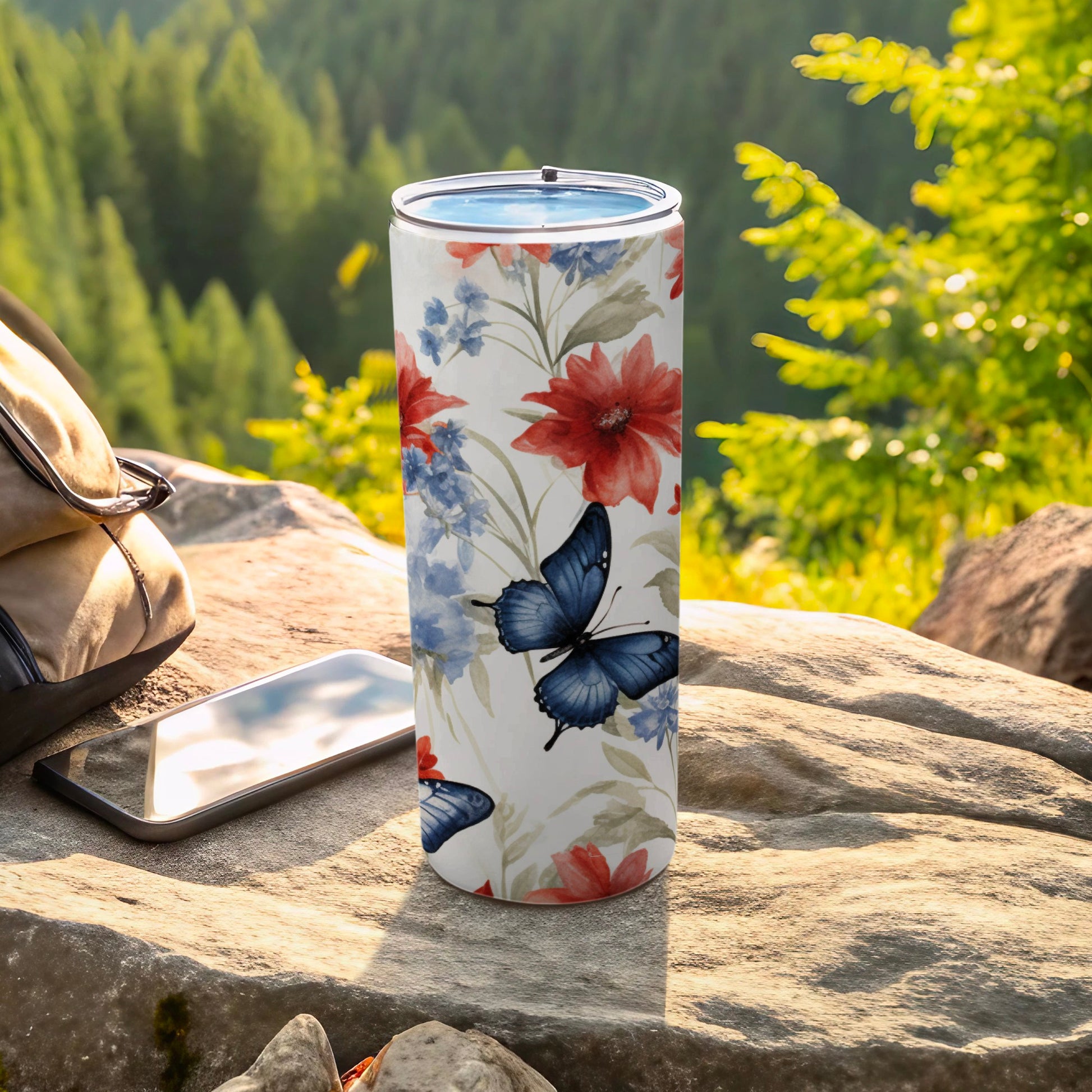 Floral and butterfly pattern tumbler on rock with phone and backpack.