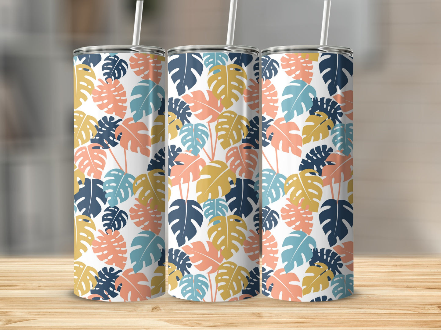 Three tropical leaf patterned tumblers with straws on a wooden surface.