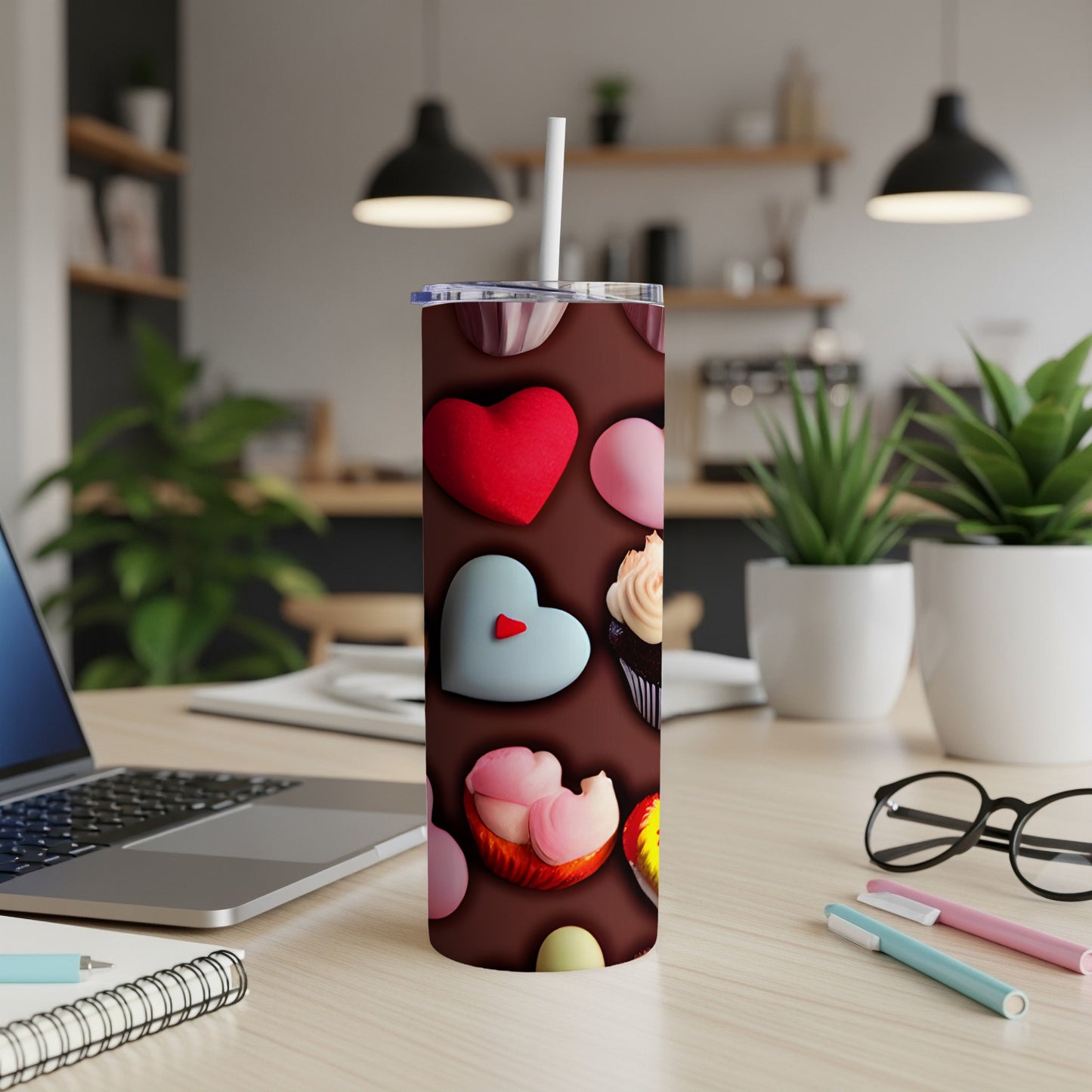 Tall tumbler with hearts and cupcakes design, straw inserted, on a desk.
