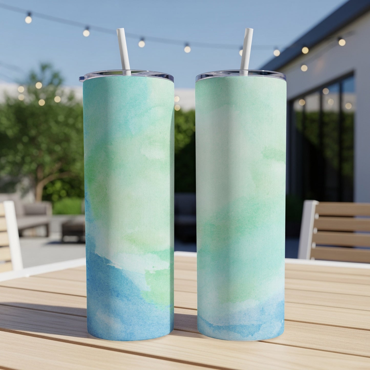 Two tall tumblers with a watercolor green and blue design sit on a wooden table outdoors.